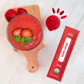 Lifestyle photo of a glass of Natierra Berry Spark Organic Smoothie on  a marble counter top with a smoothie stick pack and fresh strawberries on the side  thumbnail