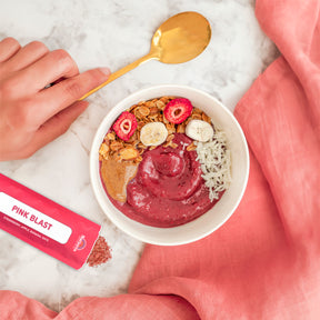 Lifestyle photo of a glass of Natierra Pink Blast Organic Smoothie on a marble counter top with a smoothie stick pack, a pink napkin and a hand holding a golden spoon on the side thumbnail