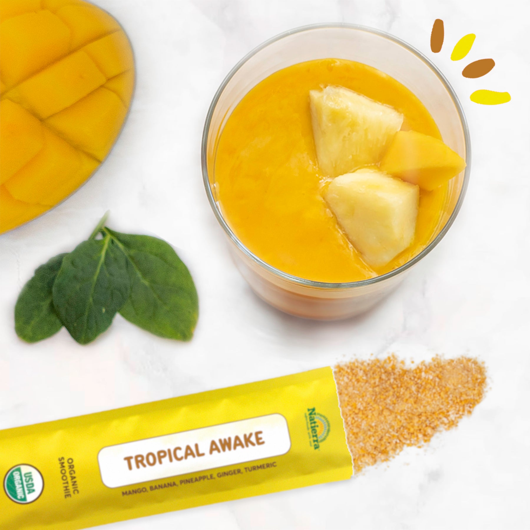 Lifestyle photo of a glass of Natierra Tropical Awake Organic Smoothie with pineapple and mango inside on a marble counter top with a smoothie stick pack, fresh mango, and spinach leaves on the side