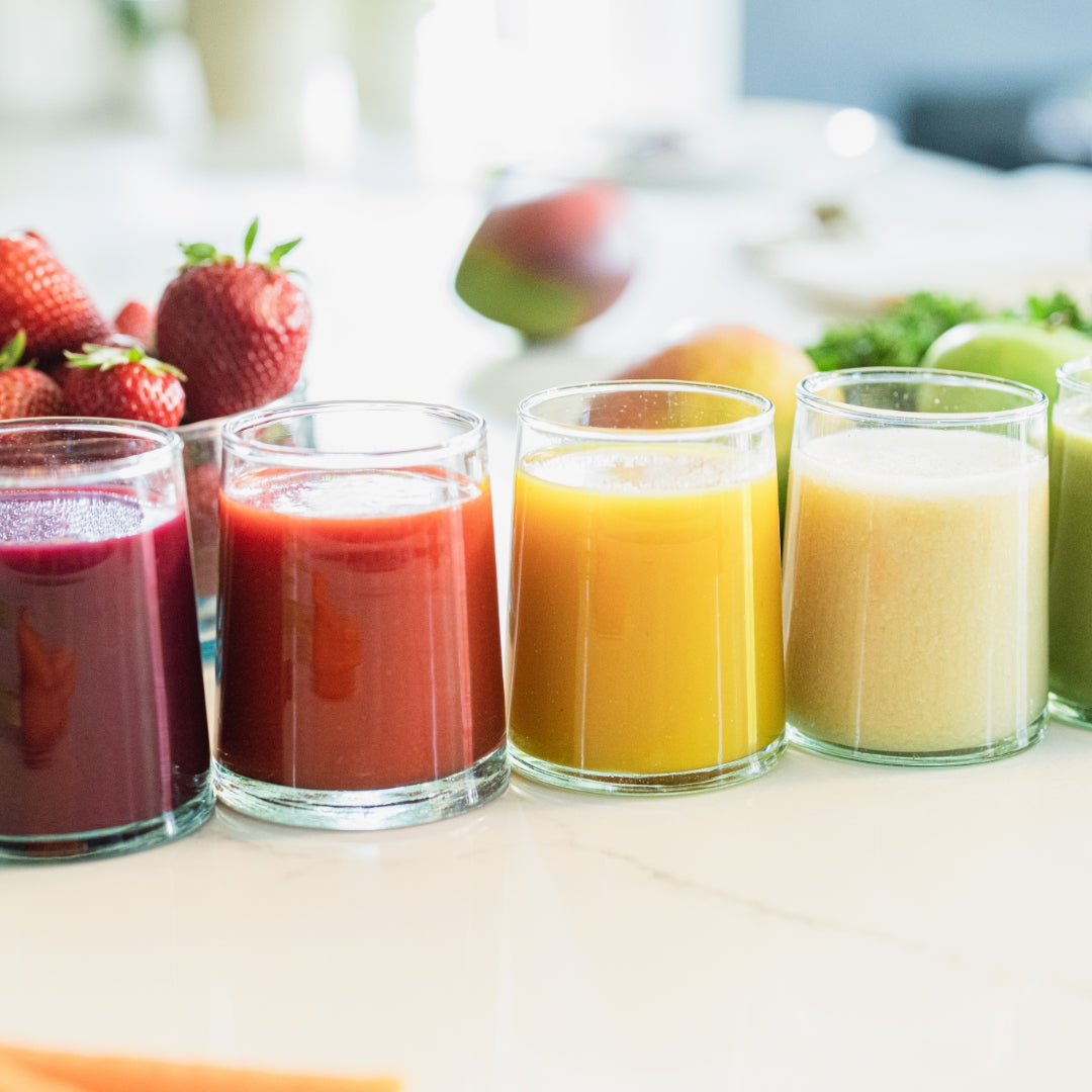 Five smoothie glasses with various smoothie colors and fruit in the background