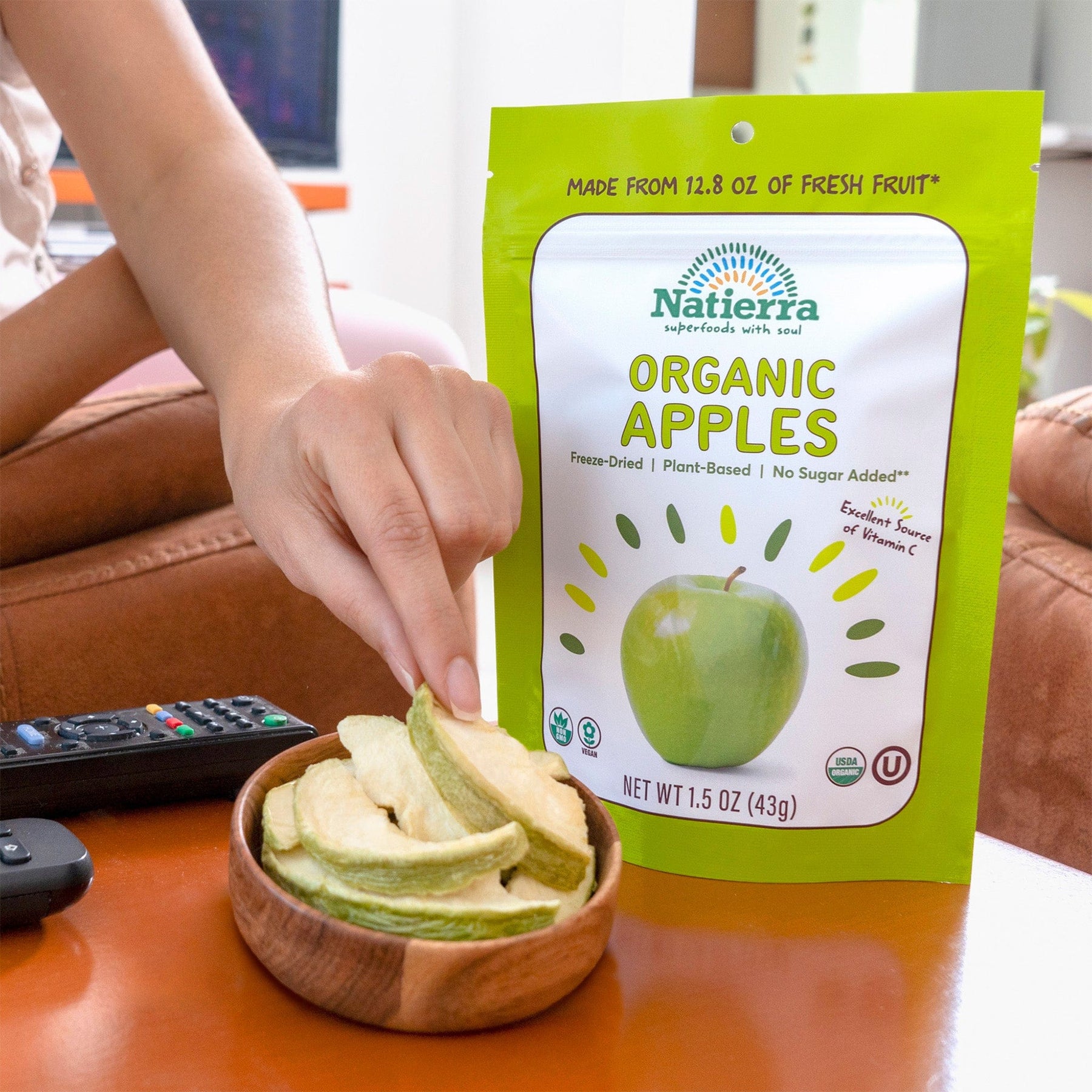 A hand grabbing apple slices from a small bowl next to a bag of Natierra Organic Freeze-Dried Apples