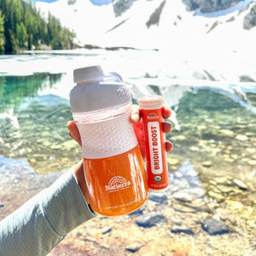 hand holding Natierra Smoothology Shaker with orange smoothie, and Bright Boost tube, lake in the background